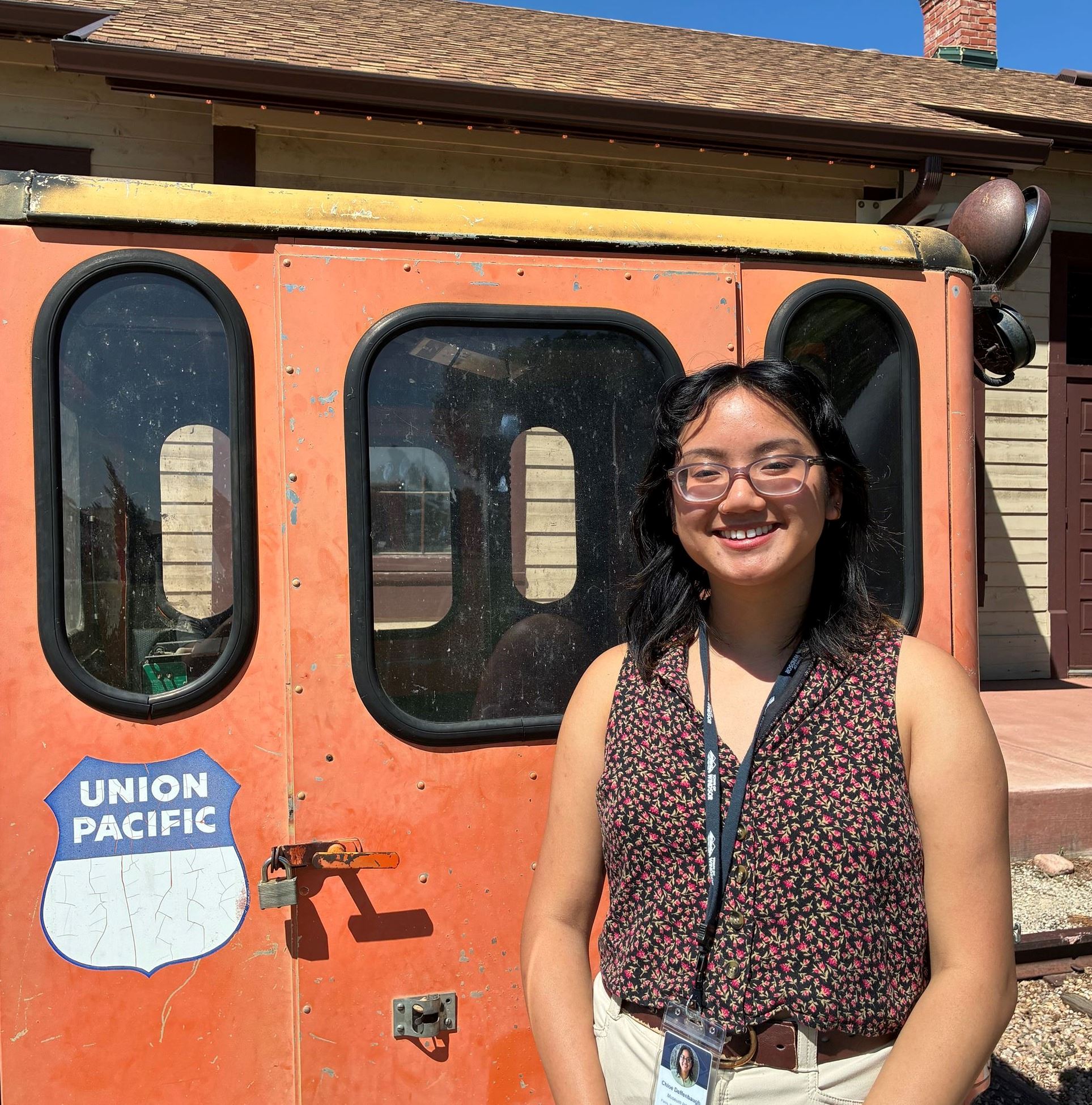 Color photograph of a girl wearing glasses standing next to an orange train car.