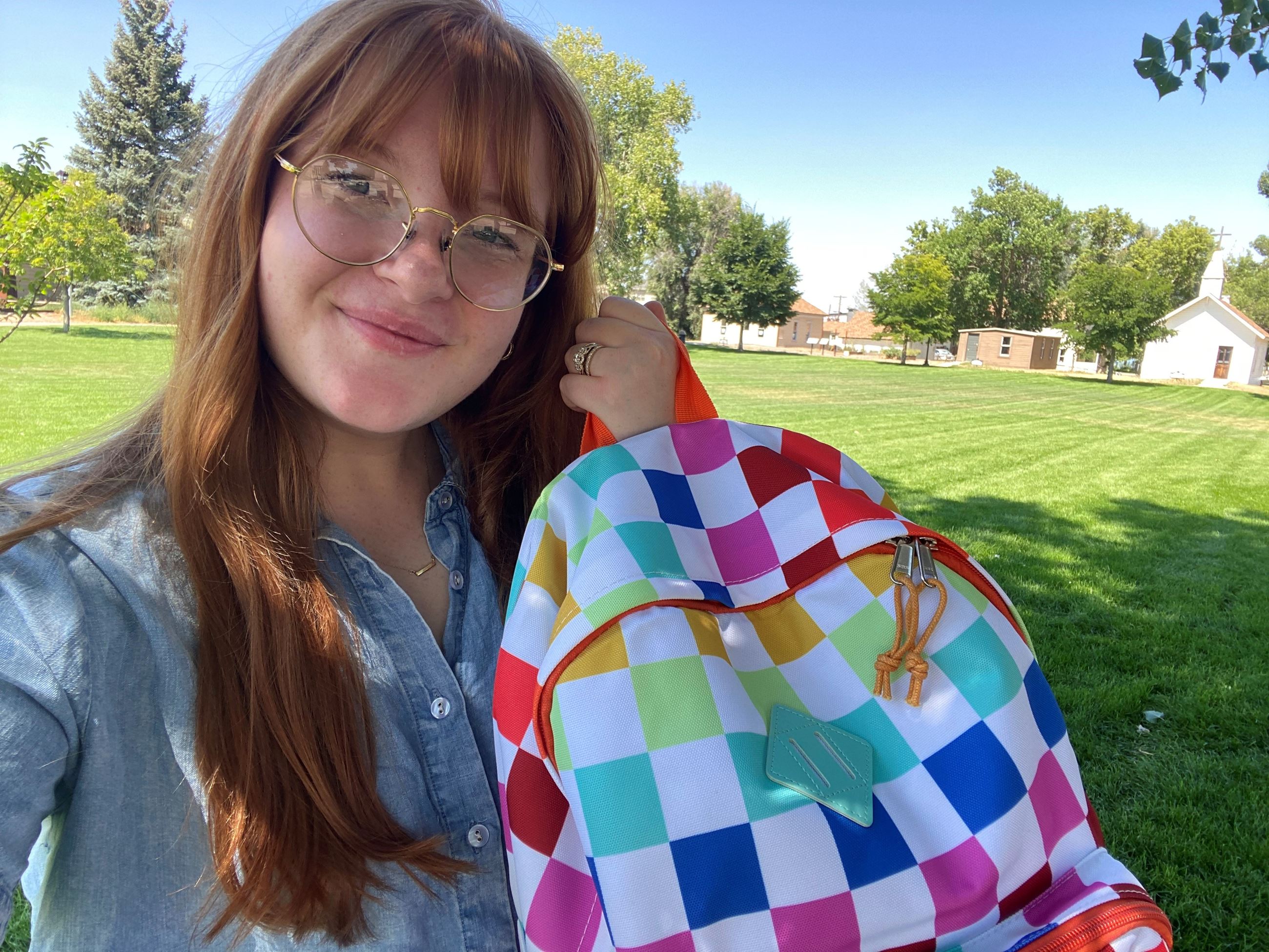 Girl with red hair and glasses holding a rainbow colored backpack.