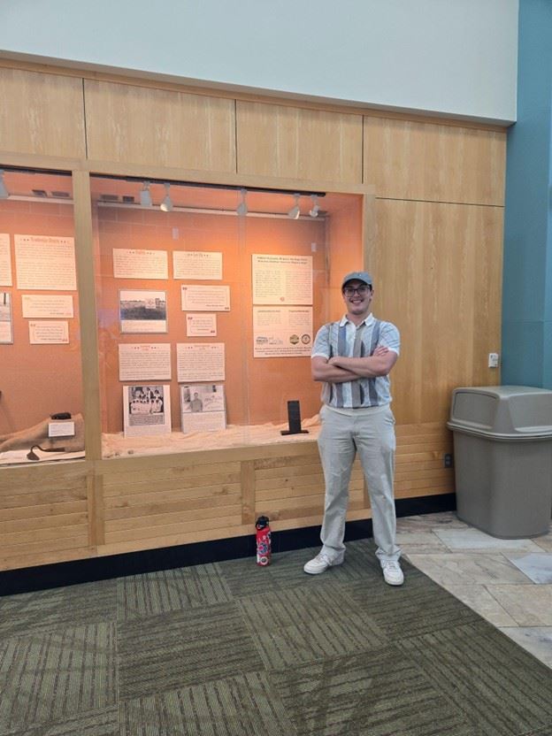 Color photograph of a young man wearing a hat standing in front of a display case.